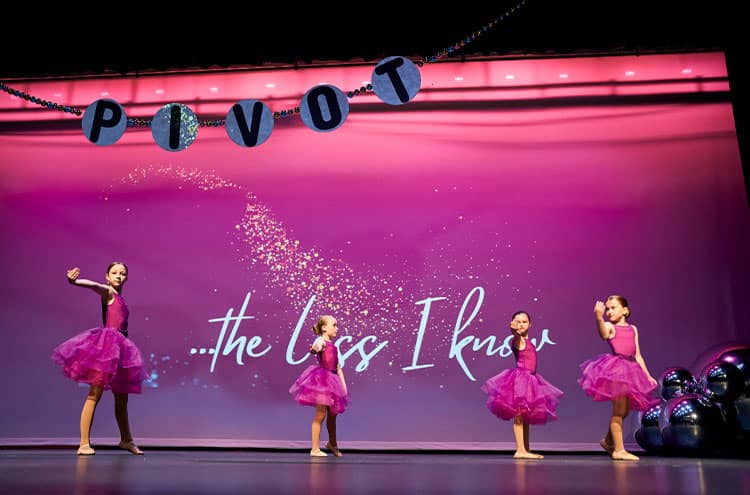 Four young girls with pink dresses are performing on stage.