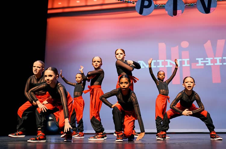 A group of young dancers in their black and orange costumes are performing on stage.