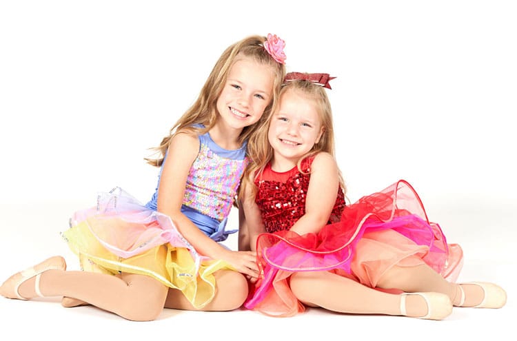 Two young dancers with their colorful dresses in all smiles and sitting down for the photo.