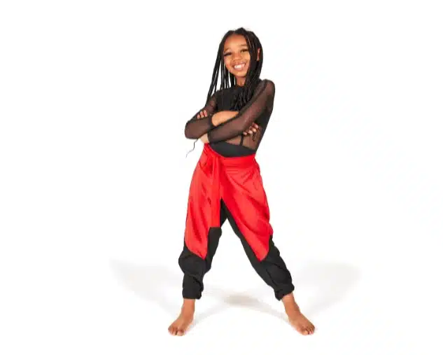 Young girl in a black mesh dance top and black pants with a red sash stands confidently with arms crossed, smiling against a white background.