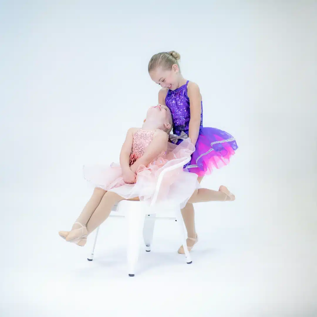 One young ballerina leans back in a chair while another stands behind her in purple and pink dancewear, both smiling joyfully.