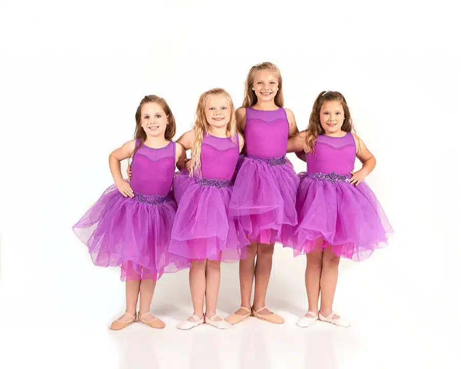 Four young girls in matching purple ballet dresses pose together with their arms around each other, smiling against a white background.