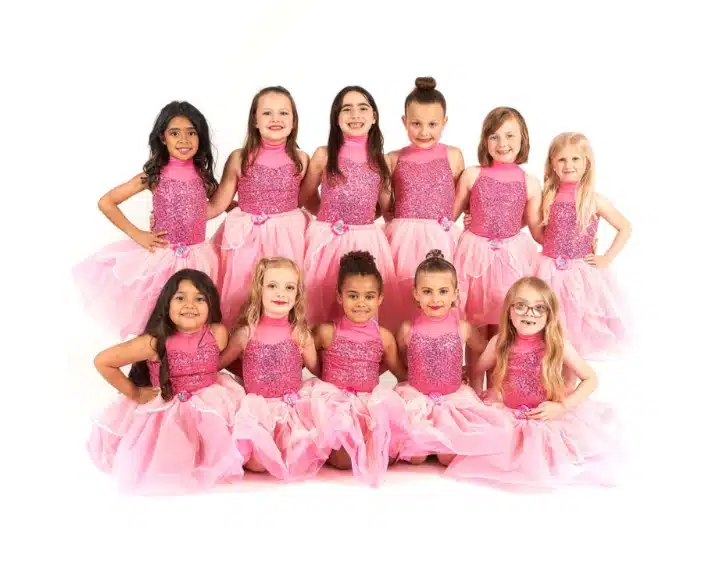 Group of young girls in matching sparkly pink dresses with tulle skirts pose together for a recital photo against a white background.