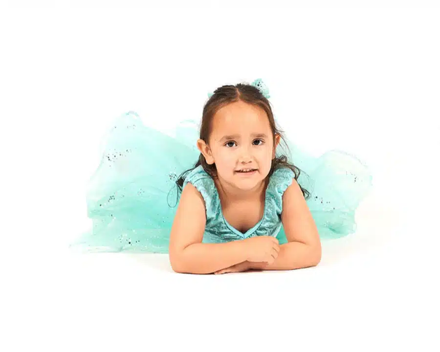 Young girl in a sparkly light blue dress lies on her stomach, smiling gently against a white background.
