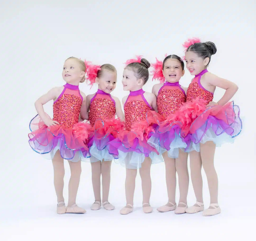 Five young girls in matching sparkly red-orange tutus pose side by side, smiling brightly.