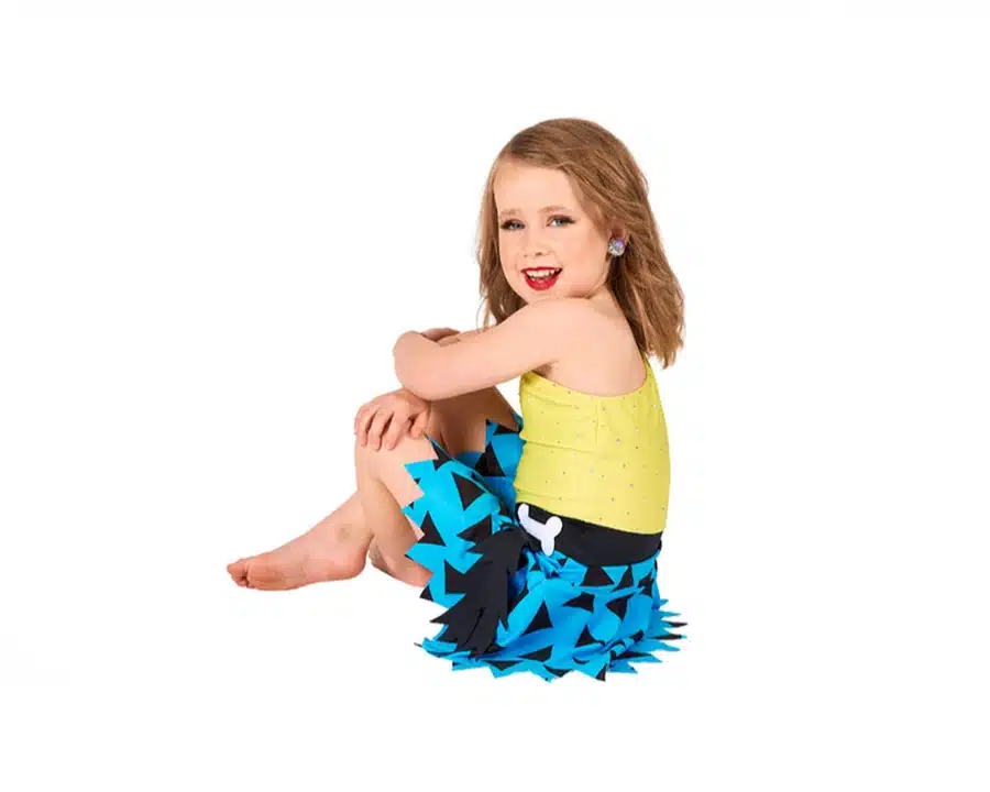 Young girl in a yellow top and blue, spiky-patterned dance skirt sits barefoot on the floor, smiling against a white background.