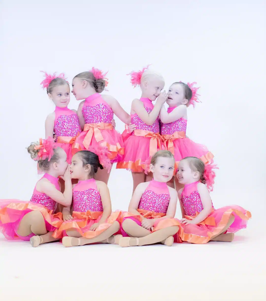 Eight young girls in matching sparkly pink dance dresses whisper and giggle in a group photo.