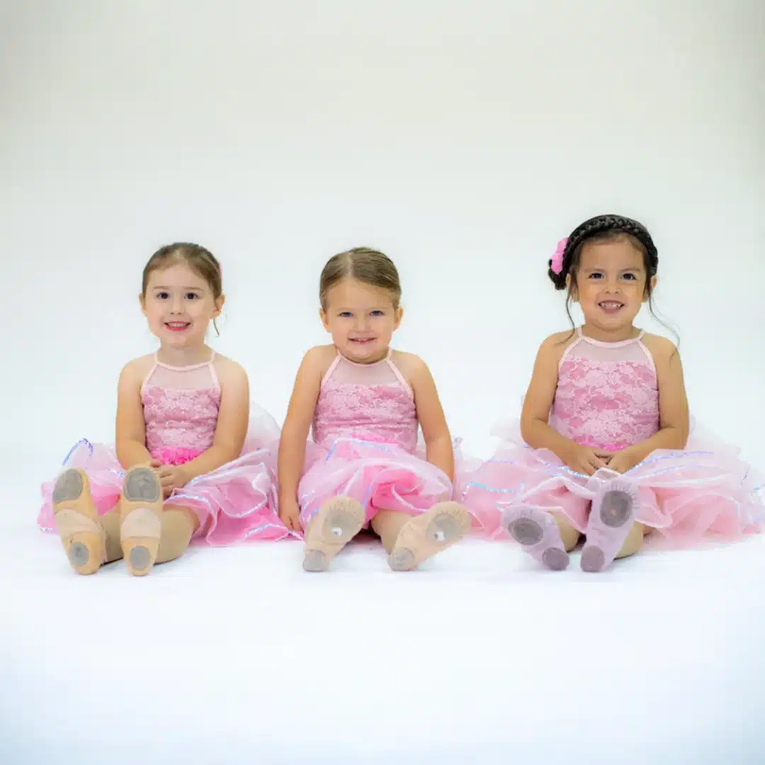 Three young girls in matching pink lace ballet dresses sit in a row on a white studio floor, smiling.