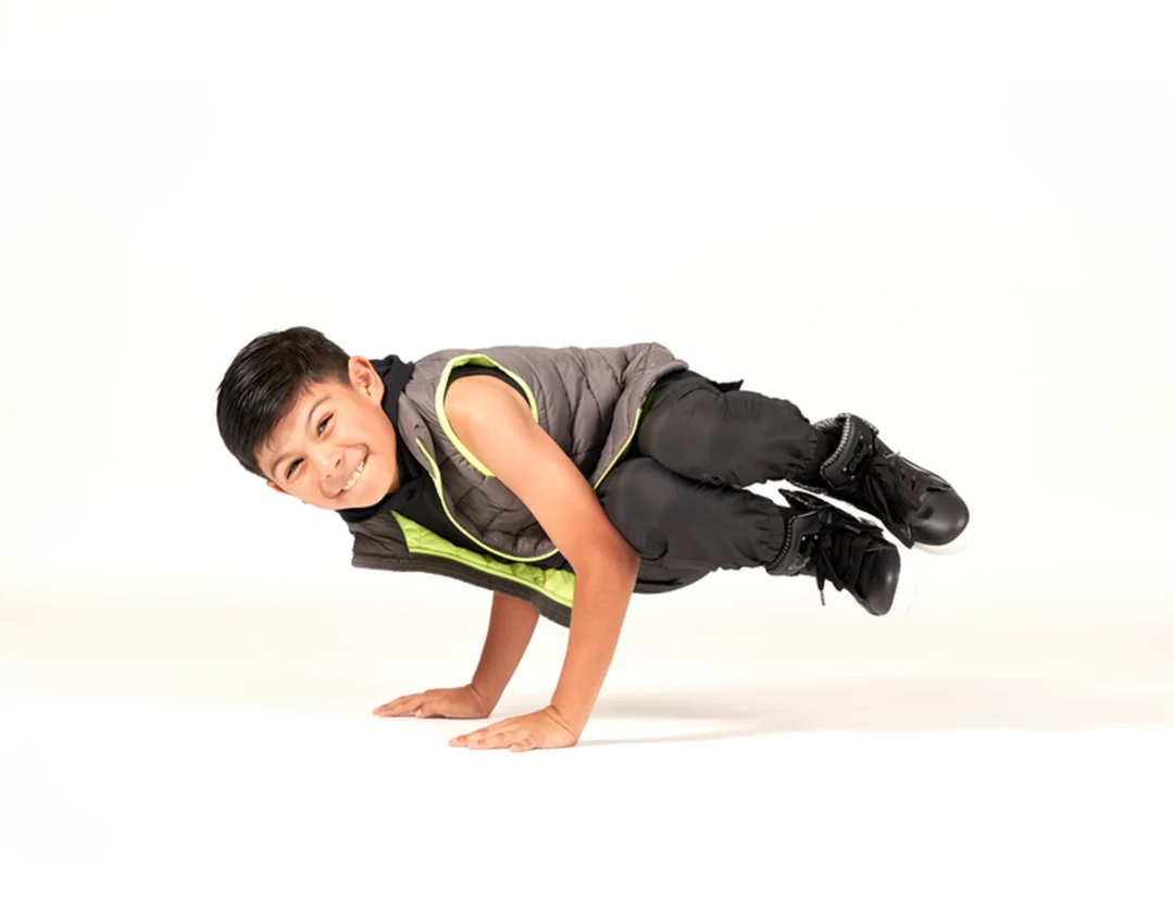 Young boy in black dancewear and a gray vest performs a breakdance freeze move, balancing on his hands and smiling against a white background.