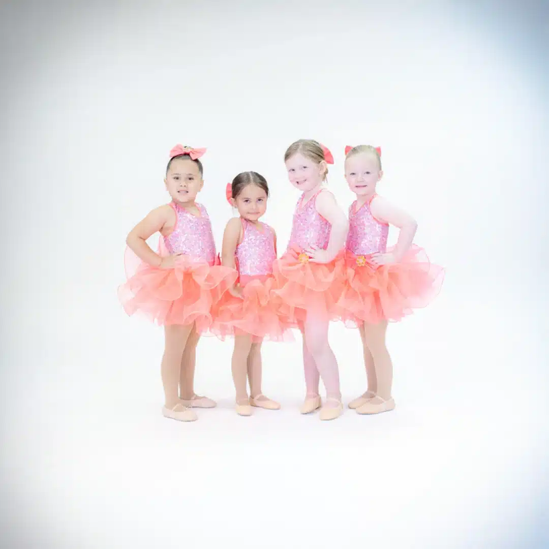 Four young girls in bright coral sequin dance dresses with ruffled tutus and bows pose together in a studio.