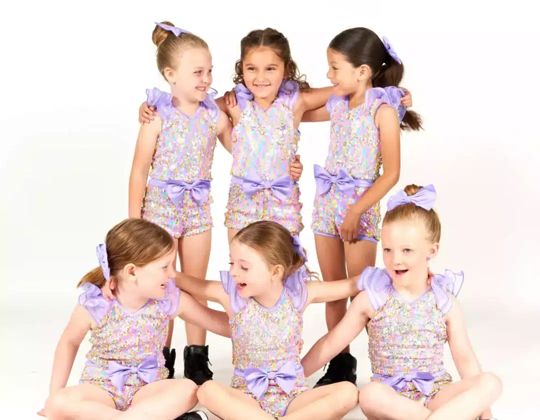Six young girls in colorful sequin rompers with lavender bows and black sneakers pose together in a playful studio group shot.