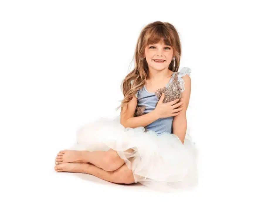 Young girl in a blue and white tutu dress sits barefoot on the floor, smiling softly against a white background.