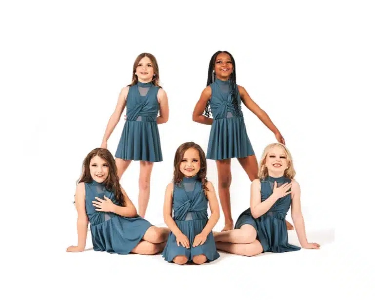 Six young girls in matching teal dance dresses pose gracefully in a studio setting, with a mix of seated and standing positions.