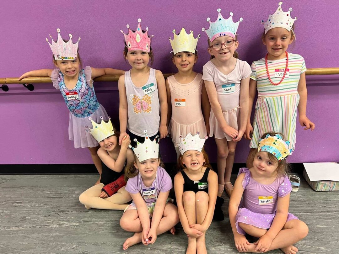 A group of young girls wearing handmade paper crowns and name tags pose happily in a dance studio with purple walls.