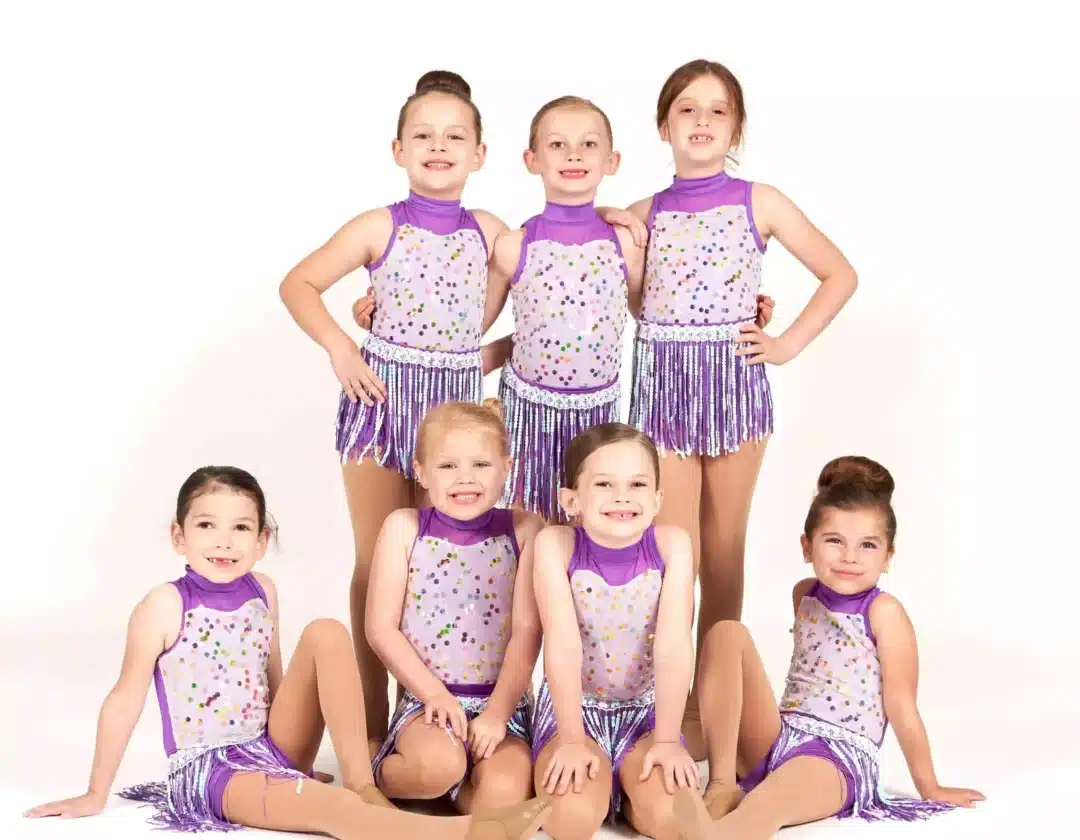 Group of young girls in purple and white sequin dance costumes with fringe skirts pose together in a studio setting.