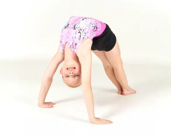 Young girl in a pink and black leotard performs a backbend, smiling upside down in a dance studio against a white background.
